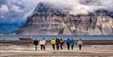 Turgruppe går på en strand. Båten, fjorden og fjell i bakgrunnen.