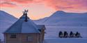 Villmarkshytte og snøskutere i snødekt landskap ved solnedgang.