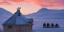 A wilderness cabin and snowmobiles in the snow landscape at sunset.