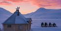 Villmarkshytte og snøskutere i snødekt landskap ved solnedgang.