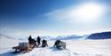Two guests and three snowmobiles in a snow-covered landscape with a clear blue sky in the background