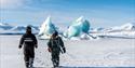 To personer går mot isfjell i snødekt landskap.
