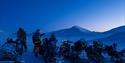 Guests and a guide standing in front of a line of snowmobiles during a break on a trip. A snow-covered landscape in blue light lies in the background.