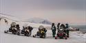 Four snowmobiles parked by the coast as a group of people gather around.