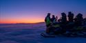 En gruppe med gjester og guide som står bak en snøscooter og ser utover et landskap med blå og oransje skumringsfarger på horisonten