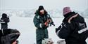 A guide preparing lunch for guests on a snowmobile trip. A guest in the background is taking photos of a glacier front in the far background.