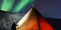 En opplyst lavvo. En person står med hodelykt utenfor. Grønt nordlys på himmelen.
