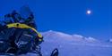 A snowmobile in a snow-covered landscape covered in blue light with a bright moon in the sky