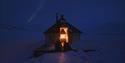Two people sitting infront of a small cabin with the door open. Inside its open fire in the fireplace. Northern lights on the sky