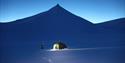 A person with a headlamp outside a tent during the blue hour, with mountain formations in the background.