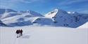 Three persons skiing in a line across a flat plateau with tall mountains in the background