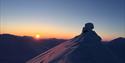 En snødekt fjelltopp i forgrunnen med en lav sol og klar himmel på horisonten over andre fjelltopper i bakgrunnen