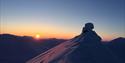 A snow-covered mountain top in the foreground with a low sun and clear skies on the horizon above other mountain tops in the background