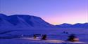 Persons on snowmobiles heading out on a trip with a mountainous landscape covered in blue light in the background