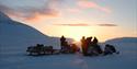 A group of guests with snowmobiles in a dark snow-covered landscape in the foreground, with a bright sky and a sunrise on the horizon in the backgroun