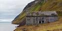 An old building on the foot of a mountain, close to the ocean with a steep mountain in the background.