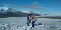 A person holding a small person on their feet on the snow with snowy mountains in the background