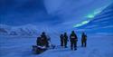 A group of people standing outside with a snowmobile. On the sky its birghr northern lights.