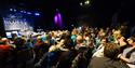 A seated audience in a concert venue with an empty illuminated stage in the background