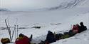 A group on a cross country skiing trip sitting down in a snow couch for a break on their way in the foreground, with an open snow-covered mountainous