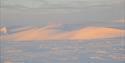 A snow-covered plain in the foreground with a snowy mountain in bright evening light in the background