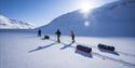 A guide and two guests pulling sleds while cross country skiing through a bright snow-covered mountainous landscape with a blue sky in the background