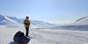 A person on skis with a sled in front of them in the foreground, with a snow-covered mountainous landscape and a blue sky in the background