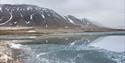 A lake with ice on its surface in the foreground with a tundra and mountainous landscape in the background