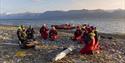 En gruppe med gjester og to guider som sitter i ring på en strand og prater sammen, med to RIB båter ankret langs sjøkanten og en fjord i bakgrunnen