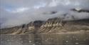 A large vast and deserted mountainous landscape with low clouds in between its mountain peaks and a fjord in the foreground