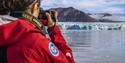 A person with a camera in the foreground taking photos across a fjord of a blue glacier front and mountains in the background