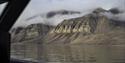 A large vast and deserted mountainous landscape with low clouds in between its mountain peaks seen through a window on board a boat