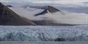 A blue glacier front along a fjord in the foreground with tall mountains surrounded by low clouds in the background