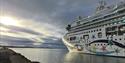 A cruise ship in port inside of a fjord, with an overcast sky and sunshine behind the clouds in the background