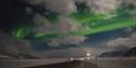 En bilvei gjennom et snødekt landskap i forgrunnen, med en mørk skyet himmel og nordlys i bakgrunnen
