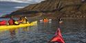 Guests paddling two and two in twin kayaks on a fjord in the foreground, with a tall mountain and a cloudy sky in the background