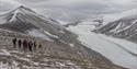 A guide and a group of guests hiking down a mountainside in the foreground with lightly snow-covered mountains and a glacier in the background