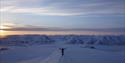 A person with outstretched arms on a snowy mountain ridge, with dark snow-covered mountains and a sunset in the background