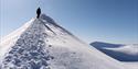 En person som går langs en fjellrygg i et snødekt landskap med en skyfri blå himmel og snødekte fjell i bakgrunnen