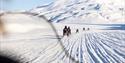 Gjester på snøscootere som kjører langs et spor i snøen i forgrunnen med en guide på snøscooter og et snødekt fjell i bakgrunnen