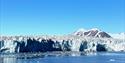 A wide glacier front with a fjord in the foreground and mountains with a clear blue sky in the background