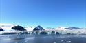 A wide glacier front with a fjord in the foreground and mountains with a clear blue sky in the background