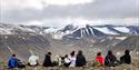 Åtte mennesker og en hund som sitter på kanten av et fjell, med en dyp dal og fjell med litt snø i bakgrunnen