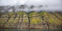 Massive green mountains rising up next to a fjord with mist in between the mountain's peaks