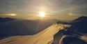 Sola som skinner på toppen av et snødekt fjell med nydelige fjell i bakgrunnen