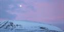 Snowmobile seen from afar in a partially snowcovered landscape, with a pink sky.