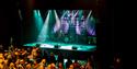 An audience mingling in a concert venue with an illuminated empty stage in the background