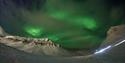 Grønt nordlys fyller himmelen over fjellene i snødekt landskap under polarnatten