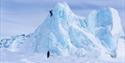A person climbing a steep ice wall while another stands at the base for safety.
