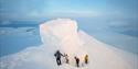 Four persons hiking along a narrow trail on top of a snow-covered mountain, with the rock formation known as Trollsteinen clad in snow behind them